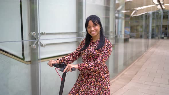 Smiling ethnic woman riding electric scooter in office building alt