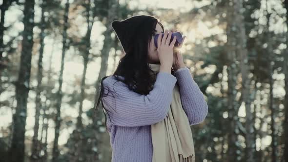 Positive Woman in Warm Hat Drinks Beverage in Winter Forest