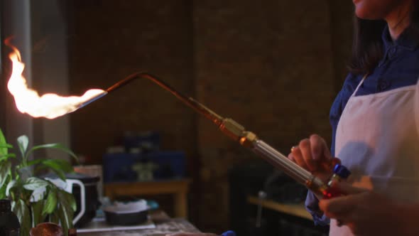 Caucasian female jeweller in workshop wearing apron and glasses, using gas burner alt