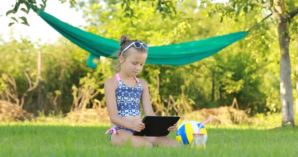 little girl in sunglasess is using laptop and lying on grass on summer sunny day. hammock and ball 