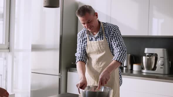 Woman Kissing Her Husband Standing at Table in Kitchen Mixing Ingredients in Bowl alt
