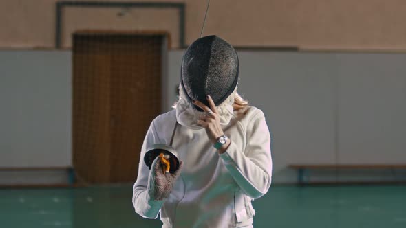 A Young Woman Fencer with Long Blonde Hair Takes Off a Protective Helmet Holding a Sword alt