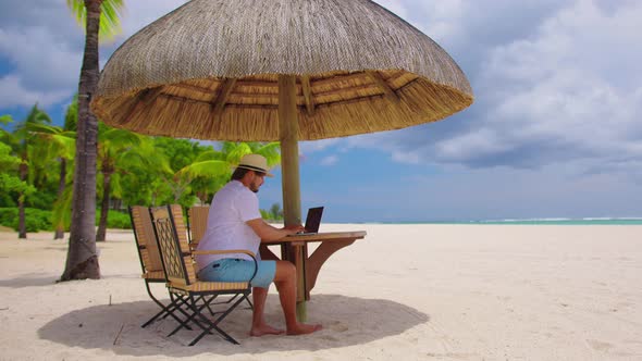 Man Relaxing with a Laptop at a Beach Resort Reclining on a Comfortable Chair Under a Straw Beach alt