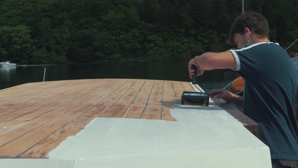 Young man applies white primer on wood boat wheelhouse roof using roller alt