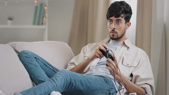 Arab Millennial Guy Bearded Indian Man Wears Glasses Lying Sofa Playing Console Enjoy Online Video alt