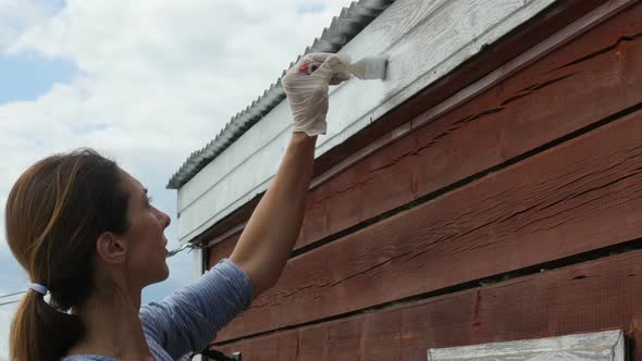 Young Woman Standing On A Stepladder And Paints A Wall Of Wooden House. Woman Makes Repairs Home
