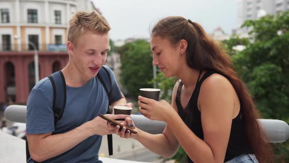 Two Young People Talking, Drinking Coffee and Using Smartphone in City alt
