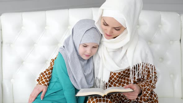 Muslim Mom in Hijab is Reading Her Little Daughter a Book Sitting on the Sofa alt