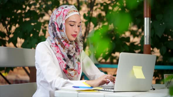 Muslim Student in Hijab Writes an Essay on a Laptop in the Park on a Summer Day alt