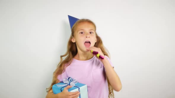 Birthday Kid Holds a Gift and Blows Noisemaker.Young Girl Wearing Birthday Cap Over White Background alt