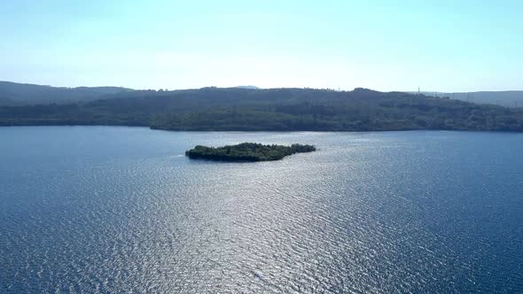 Small island with green trees in the middle of the lake with the pine forests in the background and alt