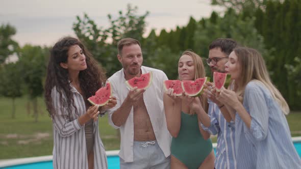 Group of young people standing by the swimming pool and eating watermelon in the house backyard