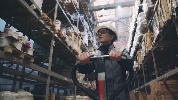 Female Worker in Hard Hat Walking Through Along Shelves with Trolley