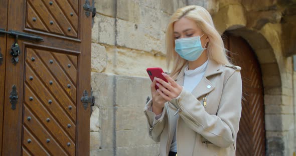 Young Woman in Protective Mask Using Smartphone in the Street of Old City. Worried Girl Searching alt