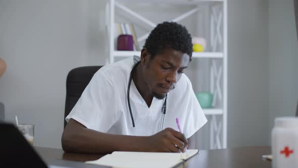 Concentrated African American Doctor Sitting at Table Writing with Pen alt