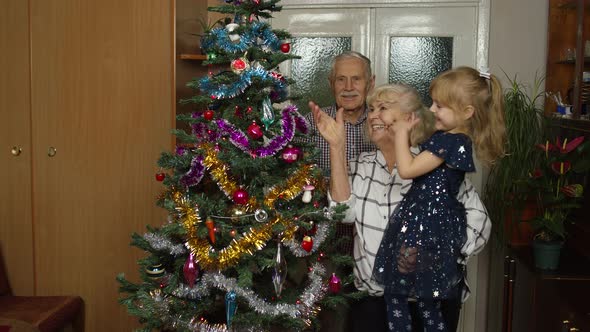 Senior Grandfather Grandmother and Granddaughter Child Lights Garlands on Artificial Christmas Tree alt