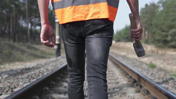 The railway worker walks along the railroad tracks, holding a sledgehammer in his hands. alt