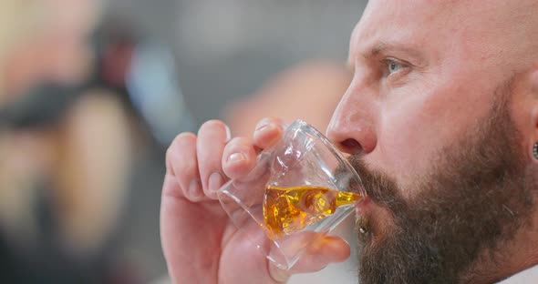 Closeup Head of Young Bearded Man with Chin Ring Drinking Whisky in Skullshaped Glass Then Improves alt
