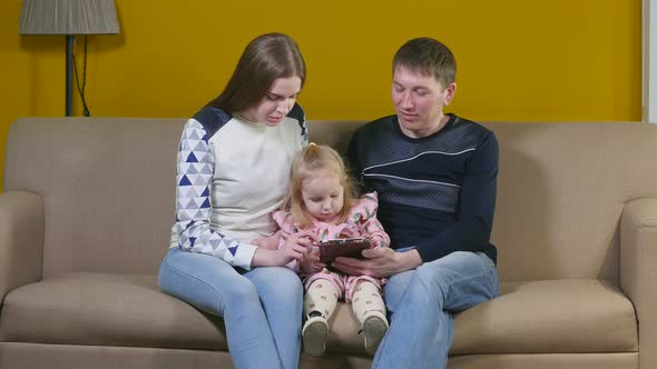 Young Parents Playing With Daughter In Tablet Sitting At Home On Couch