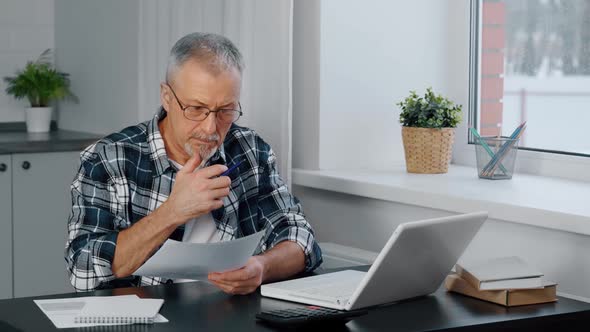 A Thoughtful Elderly Man at a Computer is Looking Through Documents