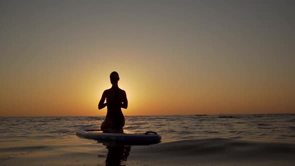 Silhouette of Young Woman Kneeling in Yoga Asana on Supboard in Slowmotion Opposite Sun While Sea is alt