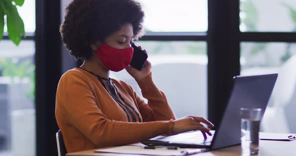 Mixed race businesswoman wearing mask sitting using a laptop in office alt