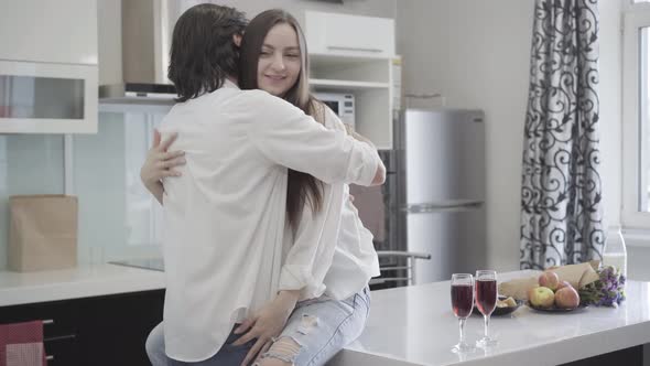 Smiling Happy Man Hugging Woman Sitting on Tabletop in Kitchen. Relaxed Young Caucasian Couple alt