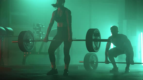 A Man and a Woman in a Colored Neon Red and Blue Light in Slow Motion Lift Heavy Barbells Over Their alt