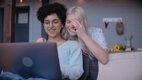 Young Girls Looks at Laptop and Laugh Girl Leans on the Sofa Hugging a Friend