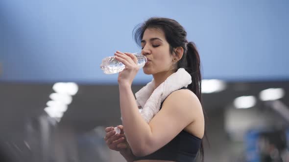 Perspiring Young Fit Middle Eastern Sportswoman Drinking Water Walking in Gym on Treadmill alt