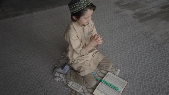 Little boy in prayer hat and arabic clothes with rosary beads and Koran book praying to God