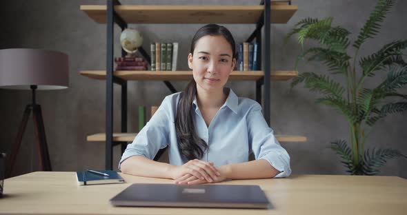 Young Asian Woman Teacher Sitting at the Table Looking at Camera and Smiling alt