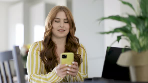 Smiling woman writing text message on smartphone while sitting at desk