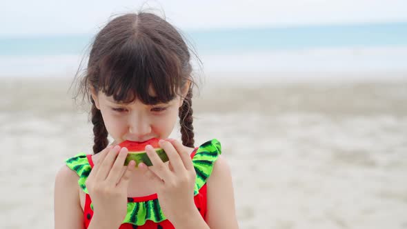 4K Little Asian girl in swimwear eating watermelon while playing on the beach in summer sunny day. alt