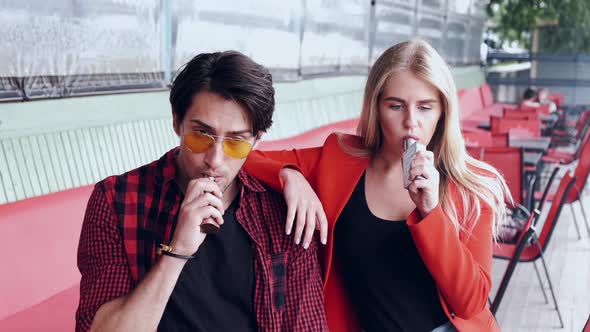Stylish Couple in a City. Two People Use the Electronic Cigarette alt