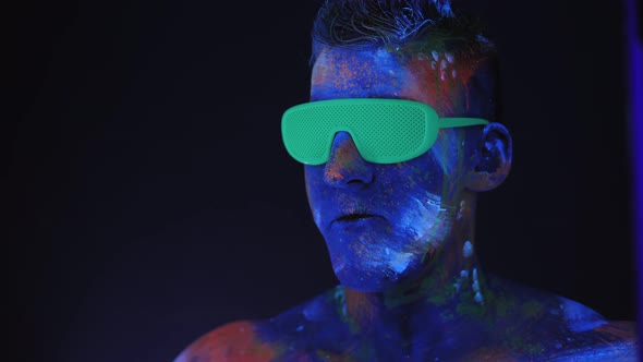 Man with UV Drawings on His Face and Neon Glasses Inflates a Bubble of Bubblegum alt