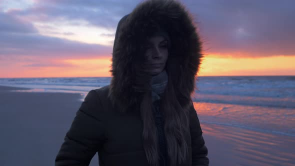 Young woman in winter clothes walks along the sandy shore of the Baltic sea beach at romantic sunset alt