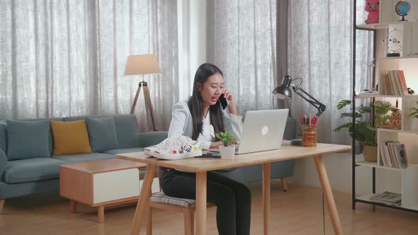 Asian Female Footwear Designer Talking On Smartphone While Working On A Laptop At Home alt