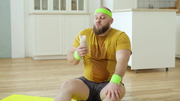 Sweaty Fat Man in Yellow Sportswear Eating Danar Sitting on Fitness Mat at Home alt