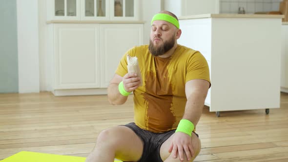 Sweaty Fat Man in Yellow Sportswear Eating Danar Sitting on Fitness Mat at Home