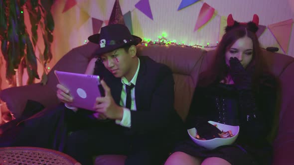 A Guy in a Creepy Costume is Actively Playing on a Tablet and the Girls are Sitting and Bored at a alt