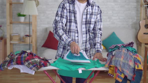 Young Asian Man Ironing Clothes on Ironing Board Looking at the Camera and Smiling alt