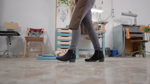 Cropped Portrait Of A Woman In Black Boots Skinny Jeans And Brown Apron Walking Inside Her Workshop alt