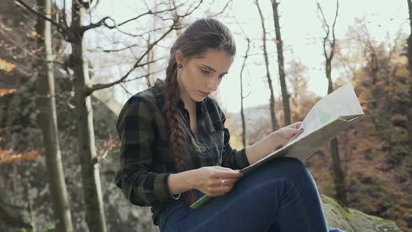 Girl with Long Hair Braided in a Braid, Sitting on a Rock in the Woods and Looking at the Map alt