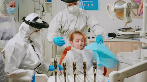 Dentist Nurse in Coverall Putting Dental Bib To Kid Patient Before Examination alt