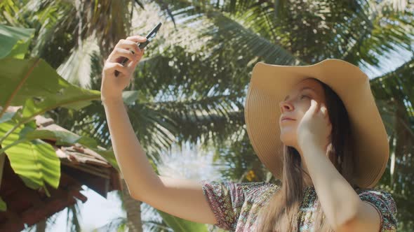 Cheerful Lady Making Selfie Photo on Mobile Phone Over Green Palm Tree Background alt
