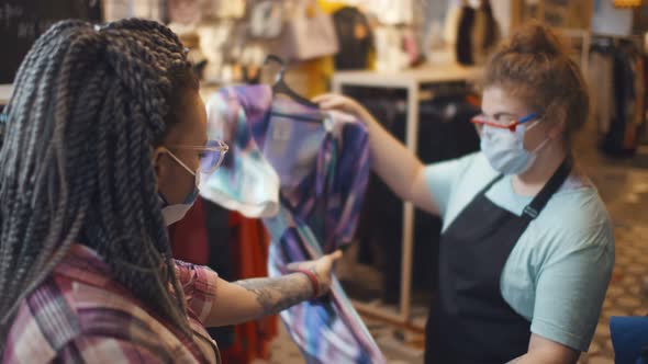 Shop Seller in Safety Mask Helping Female Customer Choosing New Dress alt