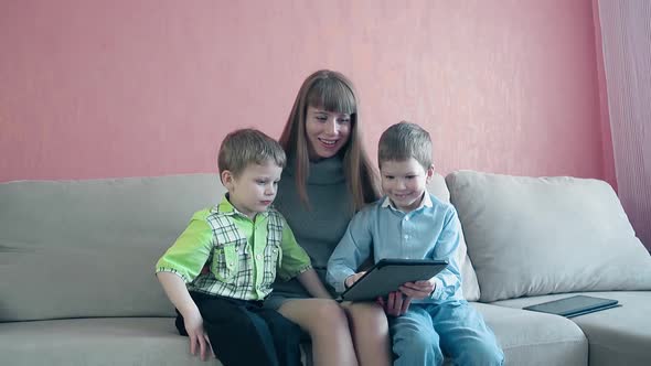 Mother and Two Little Toddler Sons Sit on Sofa Playing on Tablet alt