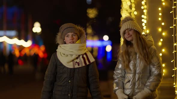 Positive Cute Boy and Girl Walking in Slow Motion with Flashing Christmas Lights at Background alt
