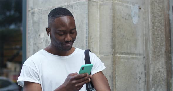 Close Up Shot of Handsome African American Guy in Casual Clothes with Earphones Using His Smartphone alt